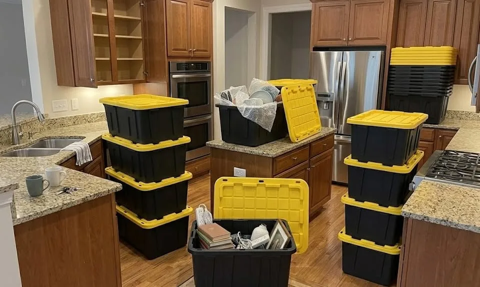 Black totes with yellow lids stacked in a kitchen, with dishes packed in open totes